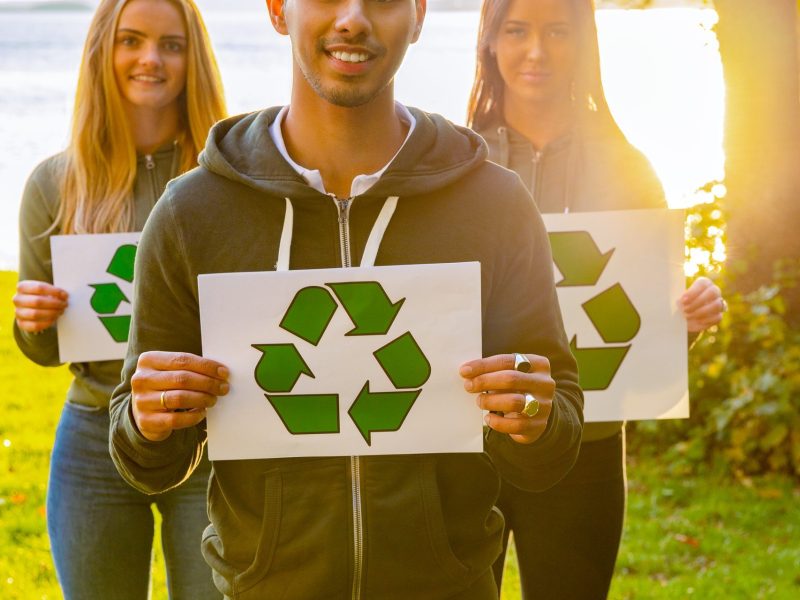 Team of young volunteers holding recycling symbol placards. Portrait of smiling man and women standing at park. They are protecting environment.